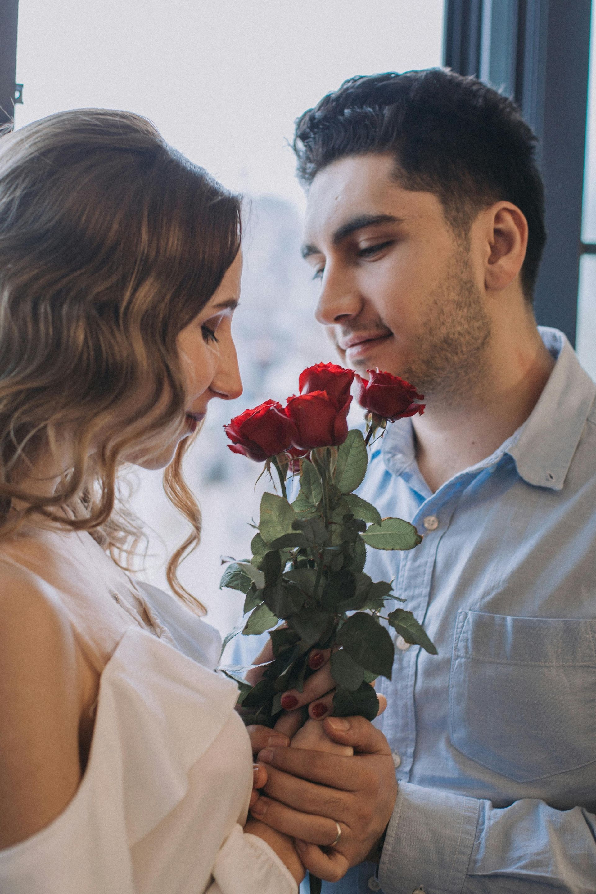 Ein Paar steht eng beieinander und schaut sich an, während es in einem sanft beleuchteten Raum einen kleinen Strauß roter Rosen hält.