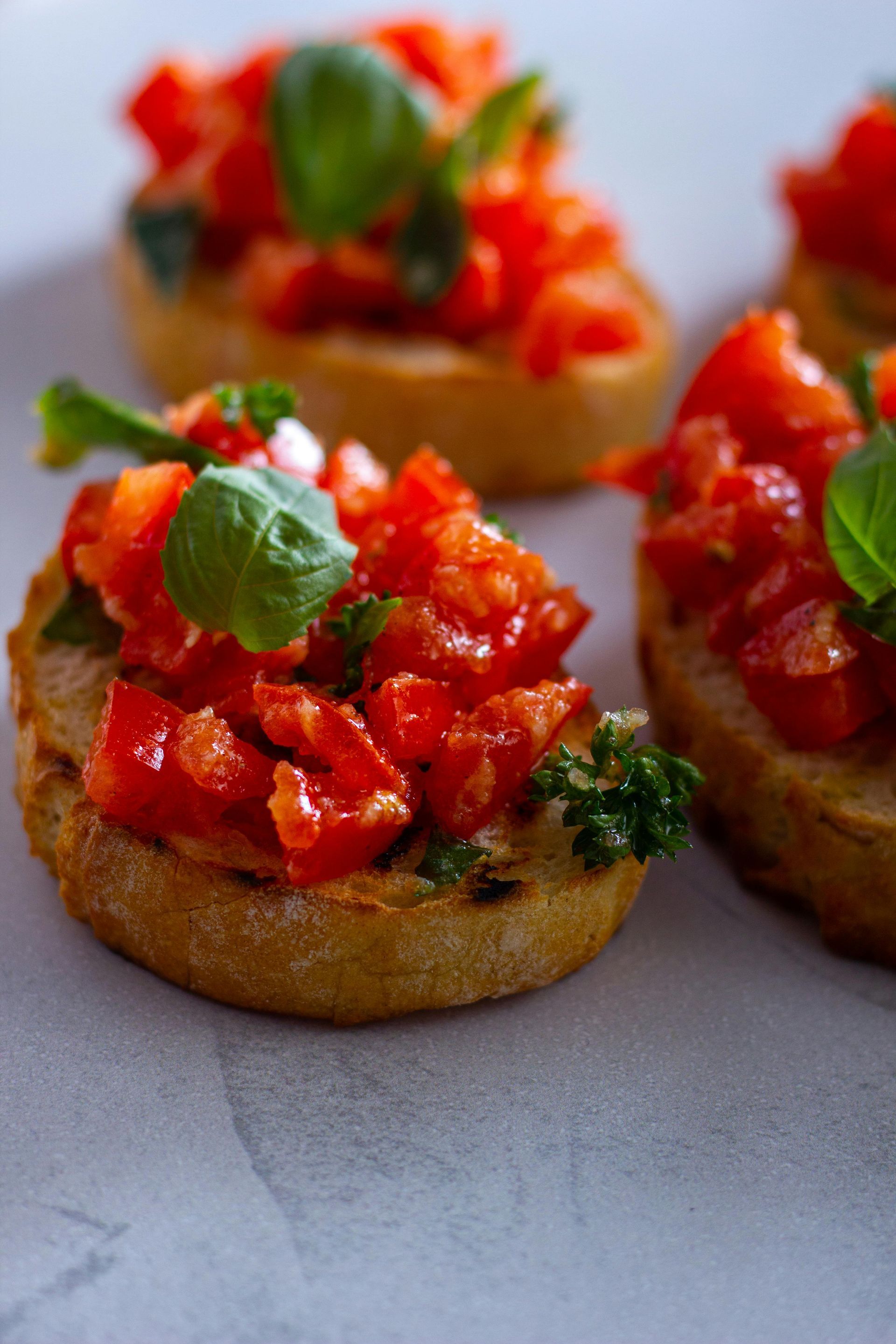 Drei Bruschetta-Toasts mit gewürfelten Tomaten, frischem Basilikum und Petersilie auf hellgrauem Untergrund.