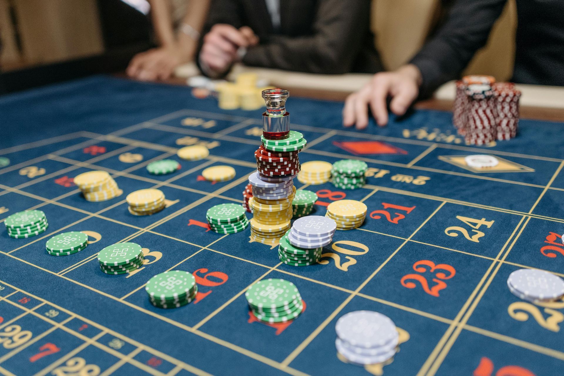Een stapel kleurrijke casinofiches op een blauwe roulettetafel; op de achtergrond zijn de vage handen van spelers te zien.