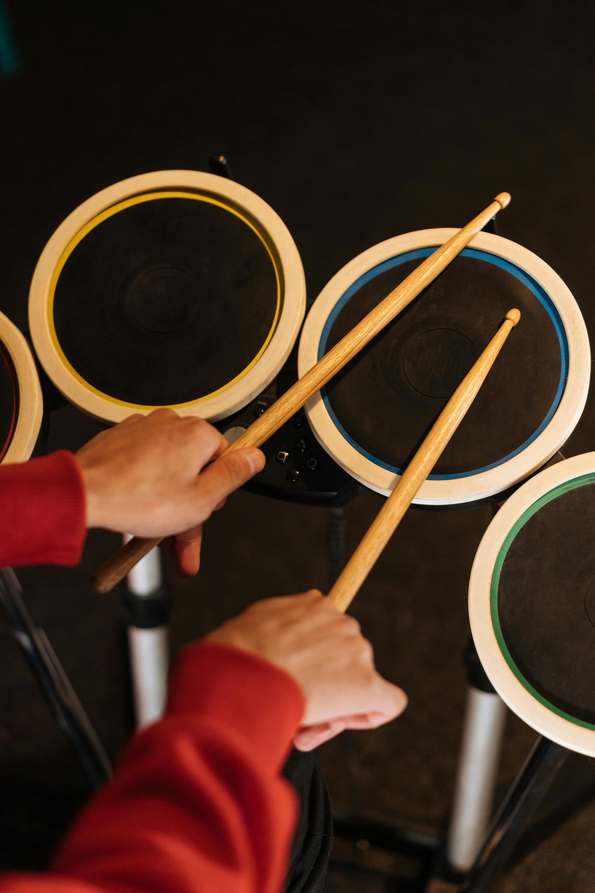 Eine Person in einem roten Pullover hält Drumsticks über elektronische Drum-Pads mit gelben, blauen und grünen Rändern.