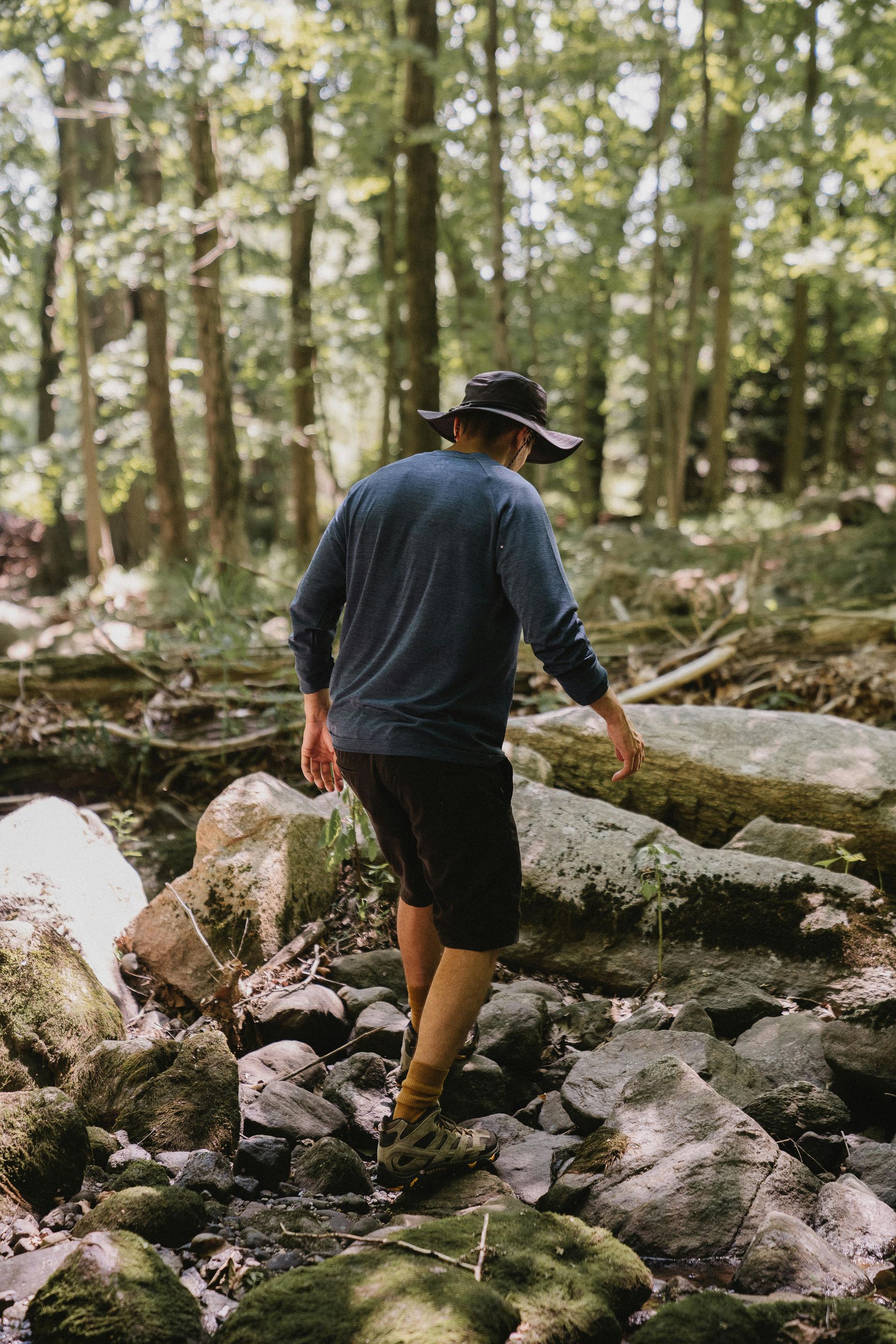 Eine Person mit breitkrempigem Hut und dunkler Kleidung schreitet vorsichtig über moosbewachsene Felsen in einem sonnenbeschienenen Wald.