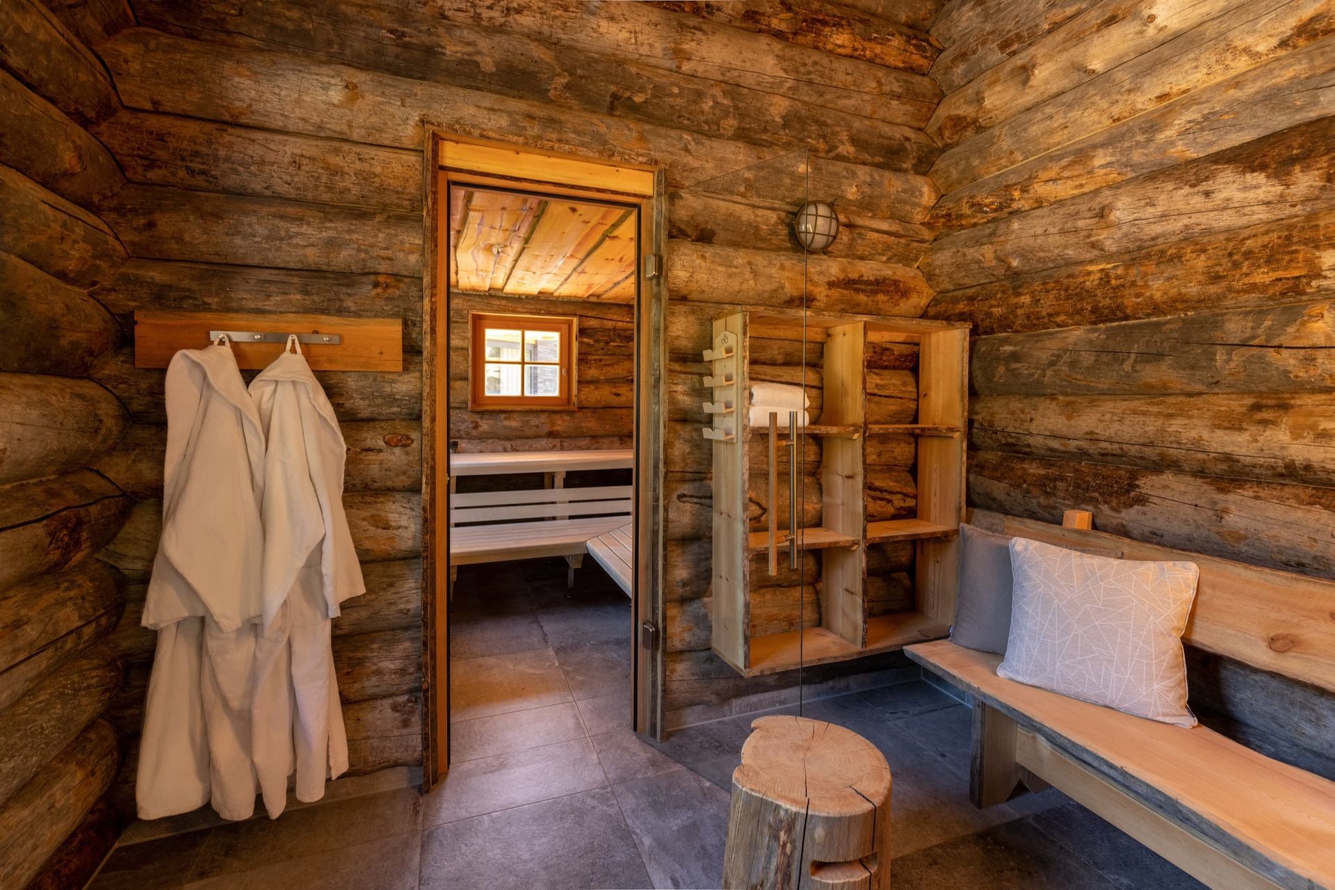 Das Interieur einer rustikalen Blockhütte zeigt weiße Bademäntel an einem Wandhaken, ein Holzregal, eine Bank mit Kissen und eine Sauna.