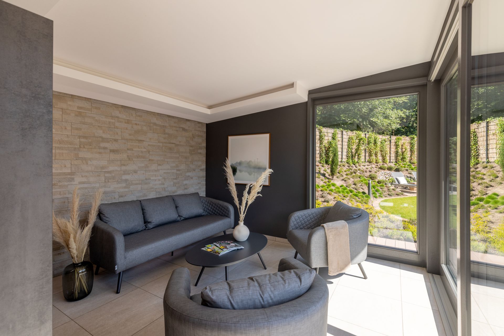 Ein moderner Wohnbereich mit einem grauen Sofa, zwei Sesseln, einem Couchtisch und einer strukturierten Wand, der sich zu einem üppigen Garten öffnet.