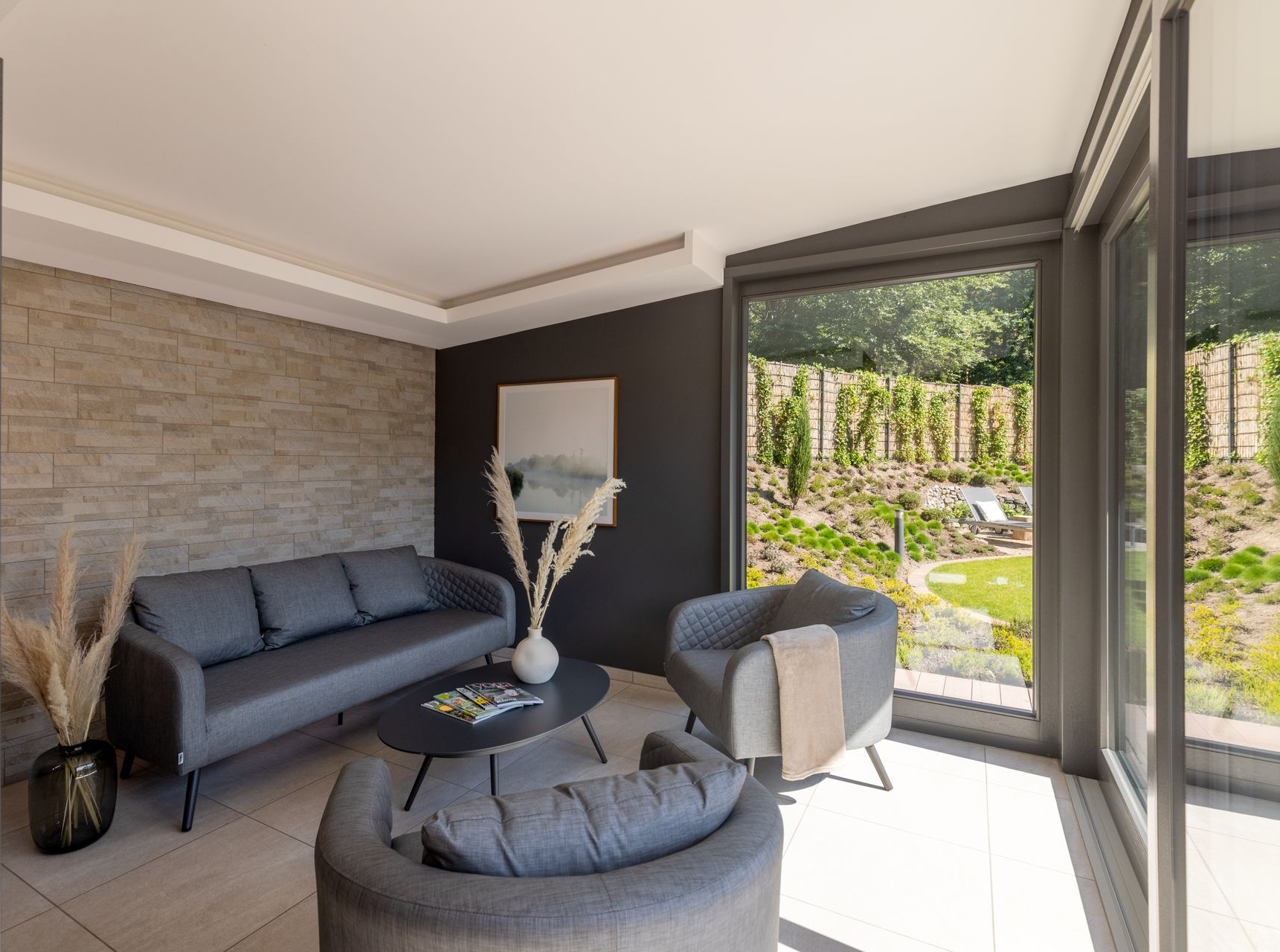 Modernes Wohnzimmer mit grauem Sofa, zwei Sesseln, einem Couchtisch und einer Wand in Steinoptik mit Blick auf einen Garten.