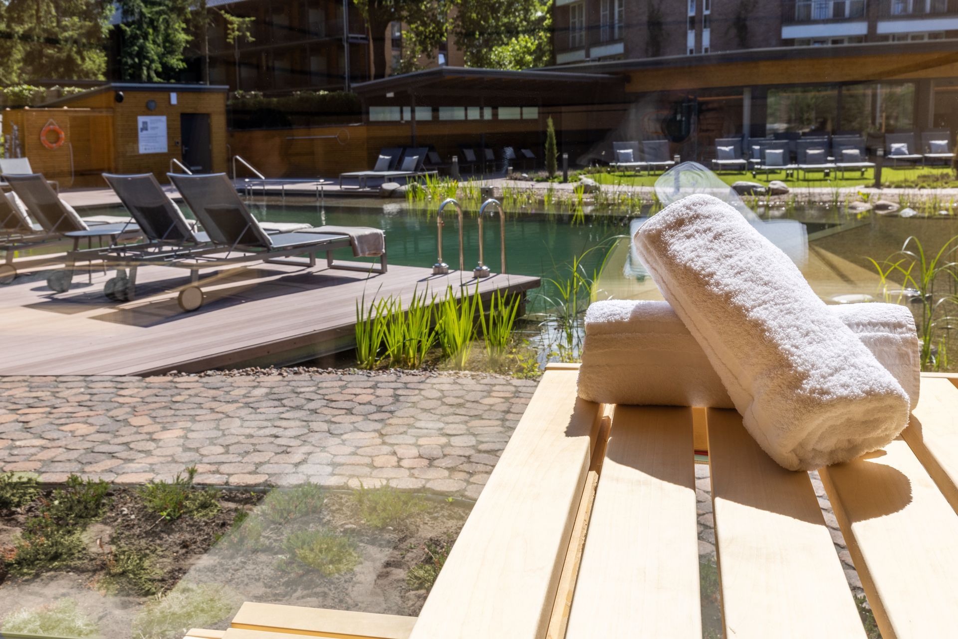 Weiße, zusammengerollte Handtücher auf einem hölzernen Liegestuhl an einem natürlichen Außenpool mit Liegestühlen auf einer Terrasse.