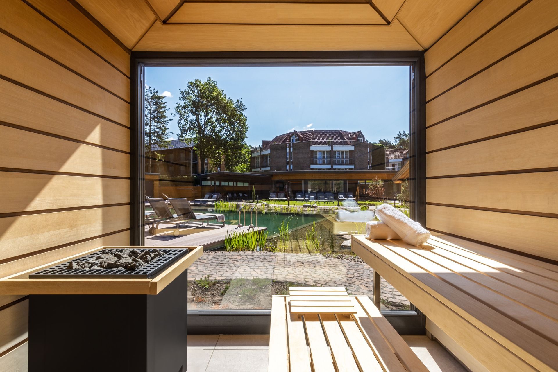 Innenansicht einer Holzsauna mit Blick durch ein großes Fenster auf einen sonnigen Außenpoolbereich.