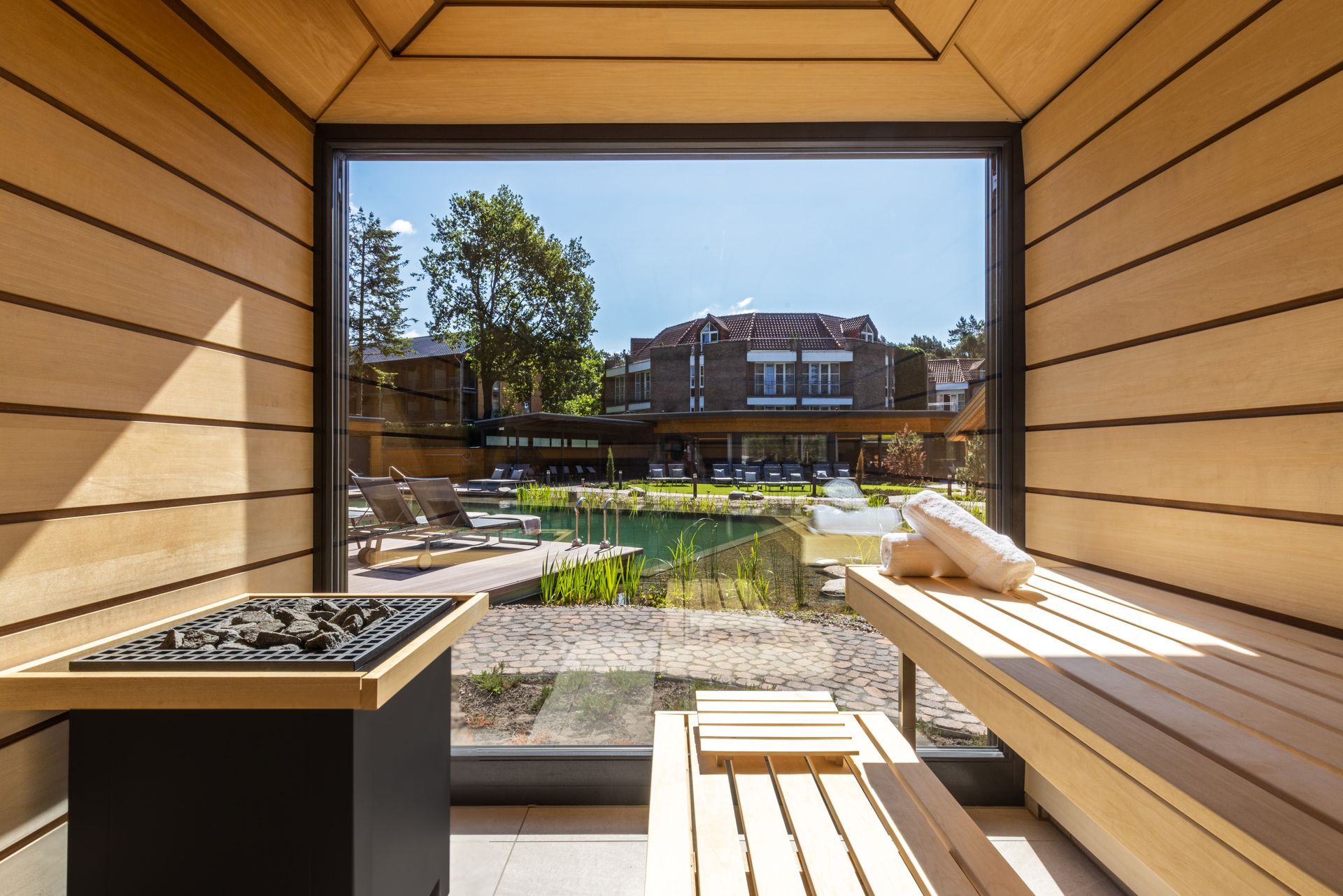 An interior view of a modern wooden sauna, looking out through a large glass window onto an outdoor pool and a terrace.