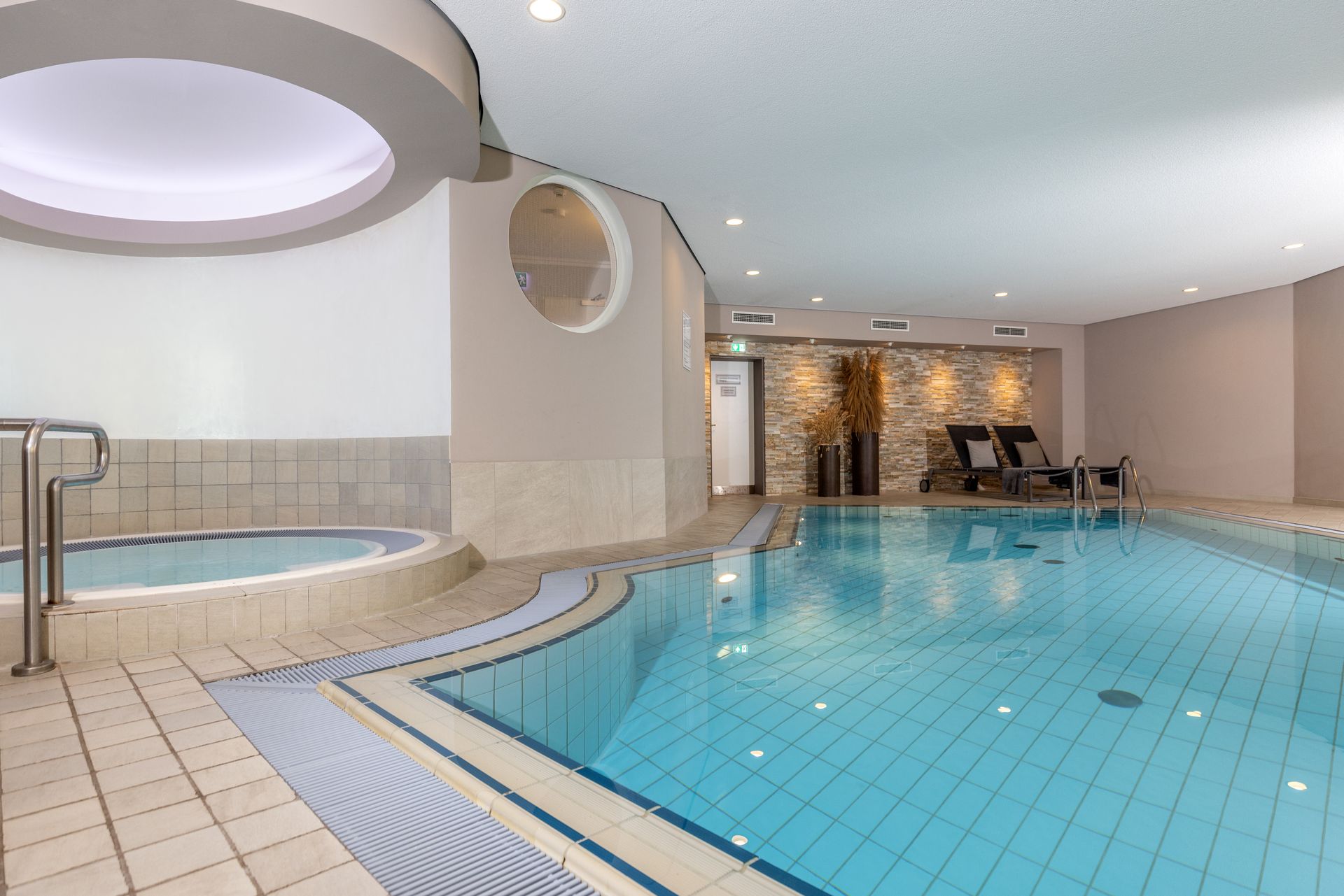 An indoor pool with a hot tub, light-colored tiles, and lounge chairs in the background.