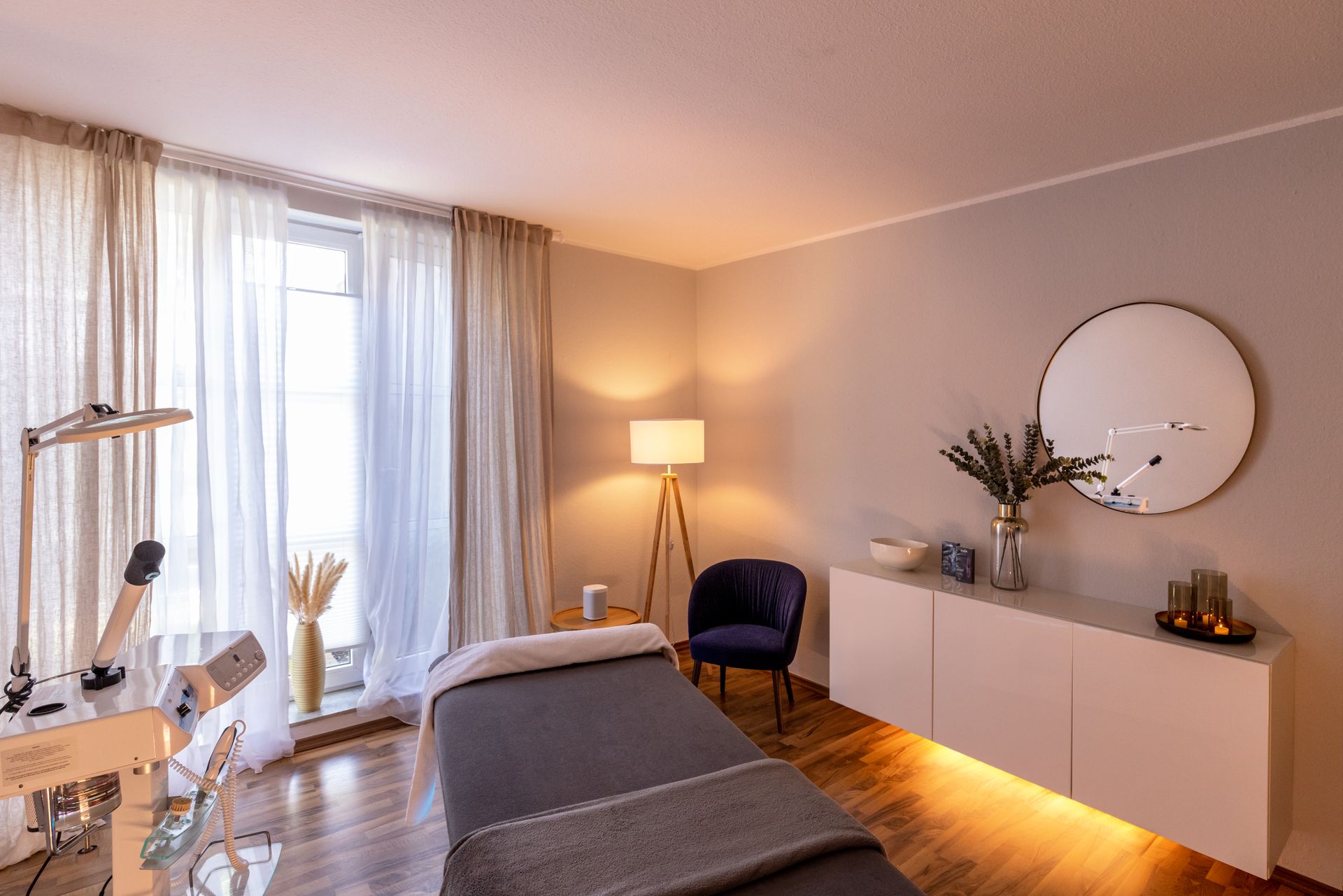 A quiet, modern massage or treatment room with a massage table, a blue velvet armchair, a floor lamp, and a white console table.