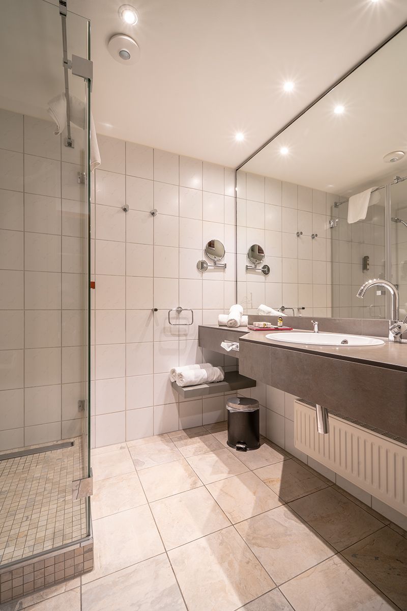 A modern hotel bathroom with a glass-enclosed shower, stone tile flooring, a vanity with a sink, and a large mirror.
