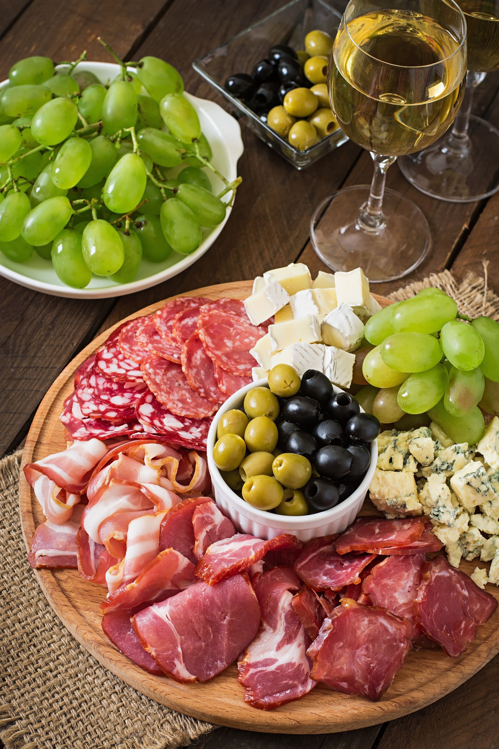 Planche de charcuterie avec raisins, fromages, charcuterie, olives et un verre de vin blanc sur une table en bois