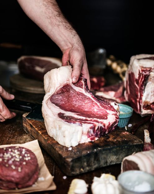Des mains découpent un gros steak cru sur un billot de boucher, avec d'autres morceaux de viande à proximité.