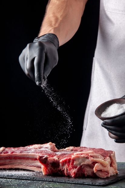 Des mains gantées assaisonnent un steak cru sur une planche à découper, le sel tombant sur la viande.
