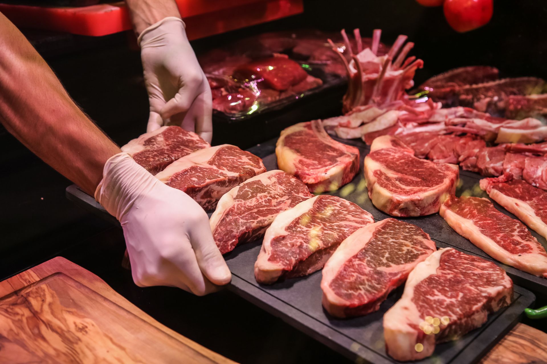 Des mains gantées disposent des steaks crus sur un plateau noir, sous une lumière rouge, devant un comptoir de boucher.