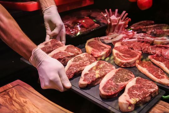 Des mains gantées disposent des steaks crus sur un plateau noir, sous une lumière rouge, devant un comptoir de boucher.