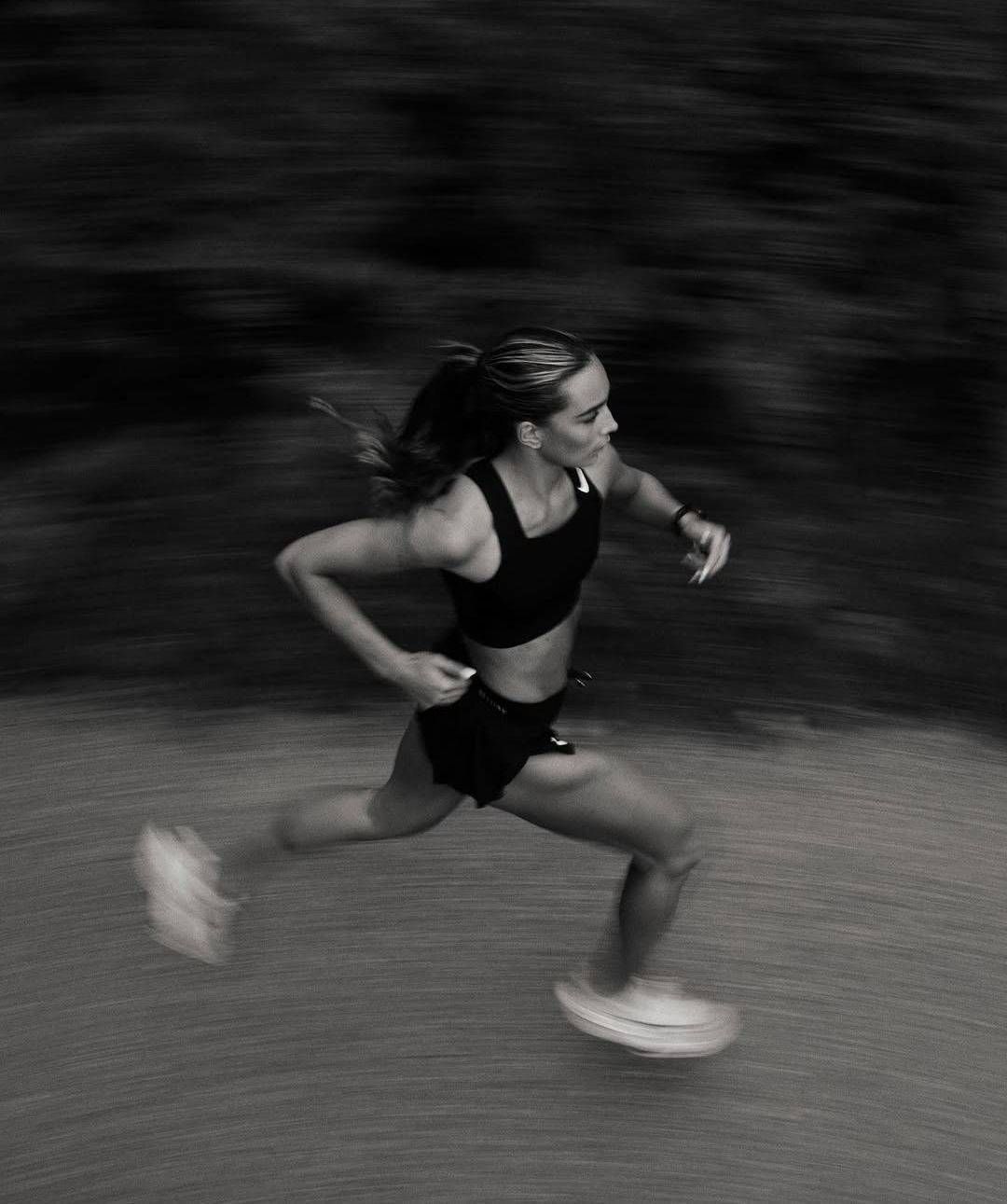 Una persona sonriente, vestida con un sujetador deportivo gris y mallas negras, corre por un puente durante el día.