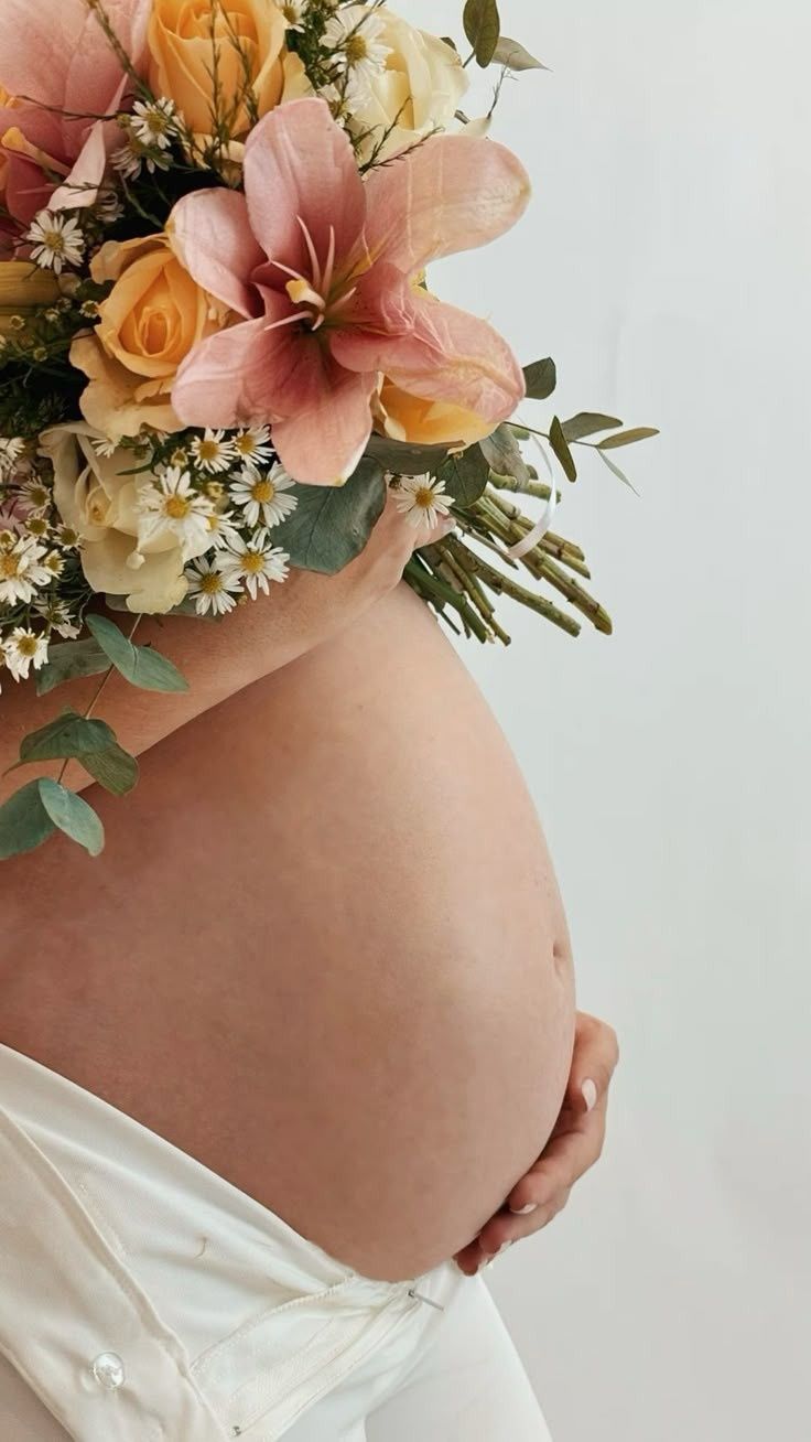 Una mujer embarazada vestida de blanco sostiene un gran ramo de lirios rosas y rosas amarillas contra su vientre.