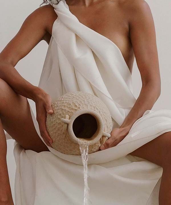 Una persona envuelta en una tela blanca sostiene un jarrón de cerámica texturizada