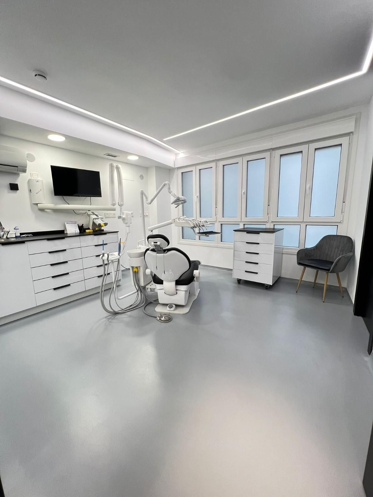 Un consultorio dental moderno y minimalista con un sillón dental blanco, gabinetes, equipo médico y un sillón gris.