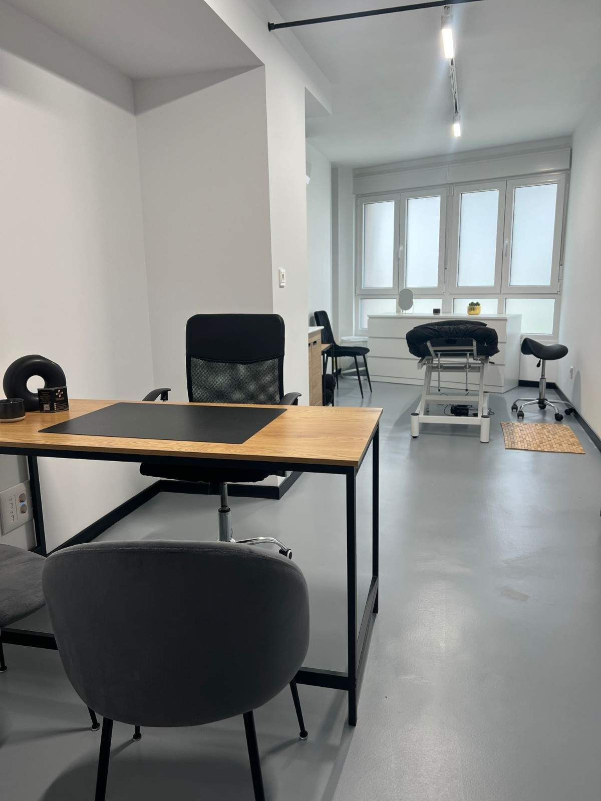 Una oficina minimalista con un escritorio de madera, dos sillas y una camilla de tratamiento de estilo médico