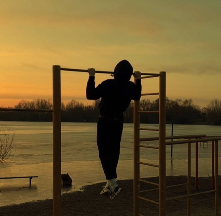 Una persona con una sudadera con capucha haciendo dominadas en una barra al aire libre, con el cielo del atardecer