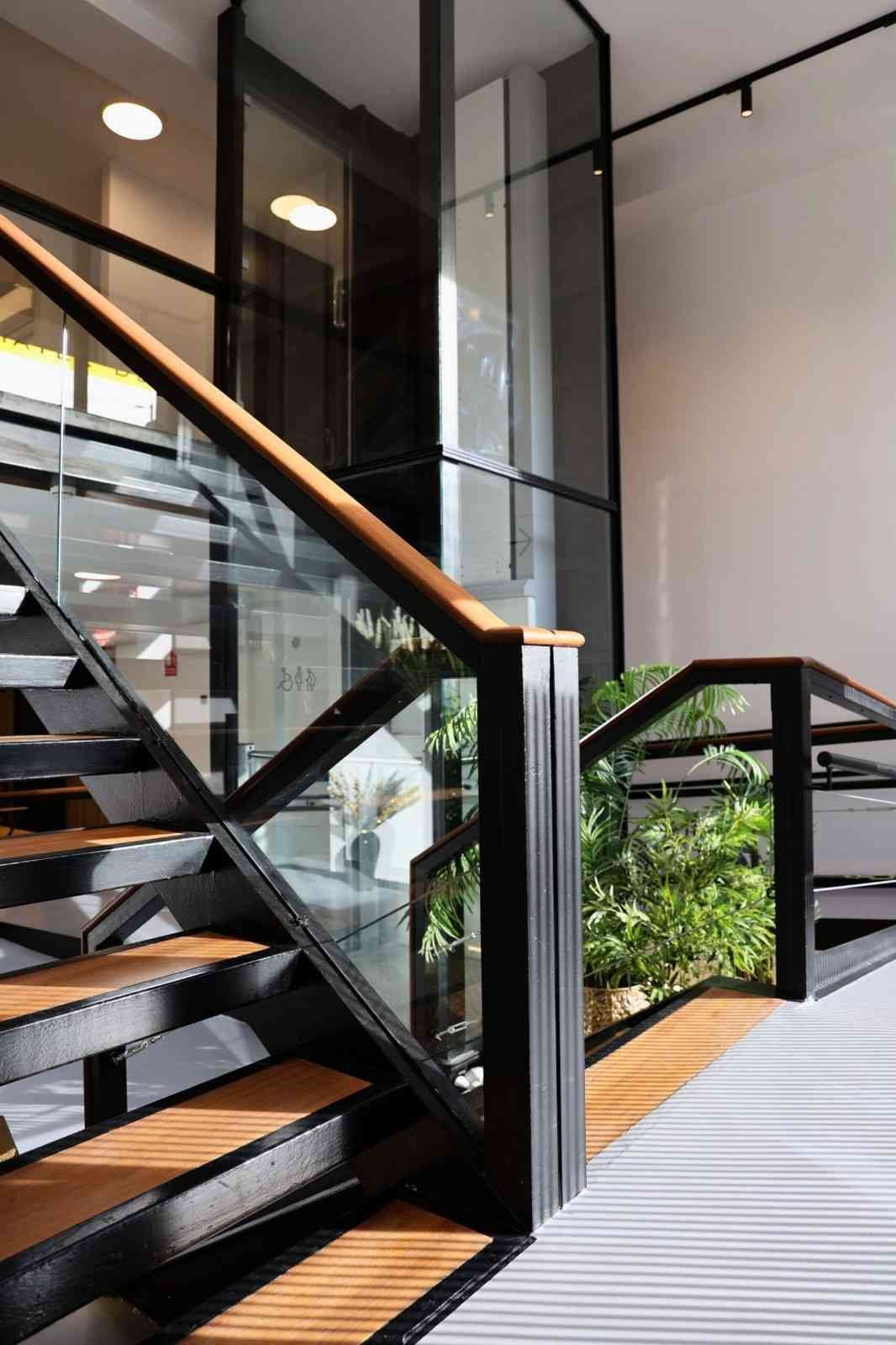 Escalera moderna con peldaños de madera, barandillas de metal negro, paneles de vidrio y un ascensor de cristal
