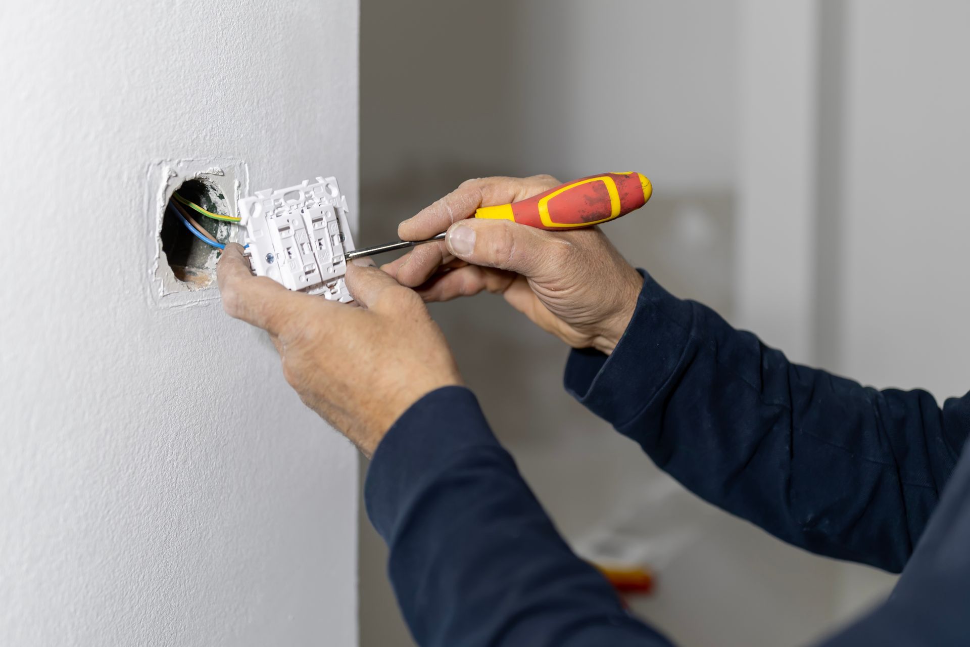 Des mains utilisent un tournevis pour installer un interrupteur électrique blanc dans un trou percé dans un mur blanc aux fils dénudés.