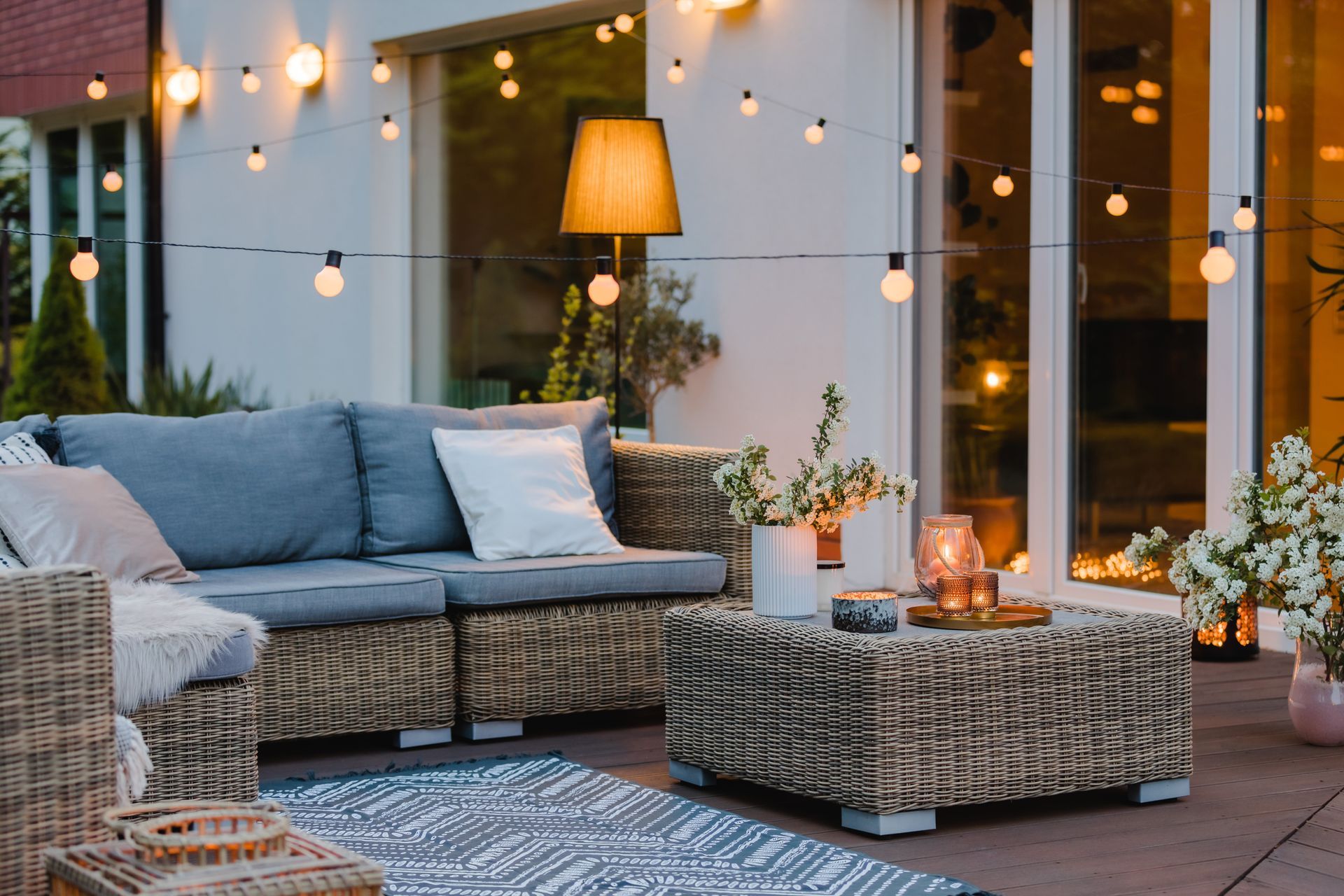 Mobilier de jardin en osier avec coussins gris et table basse sur une terrasse, illuminée par des guirlandes lumineuses suspendues au crépuscule.