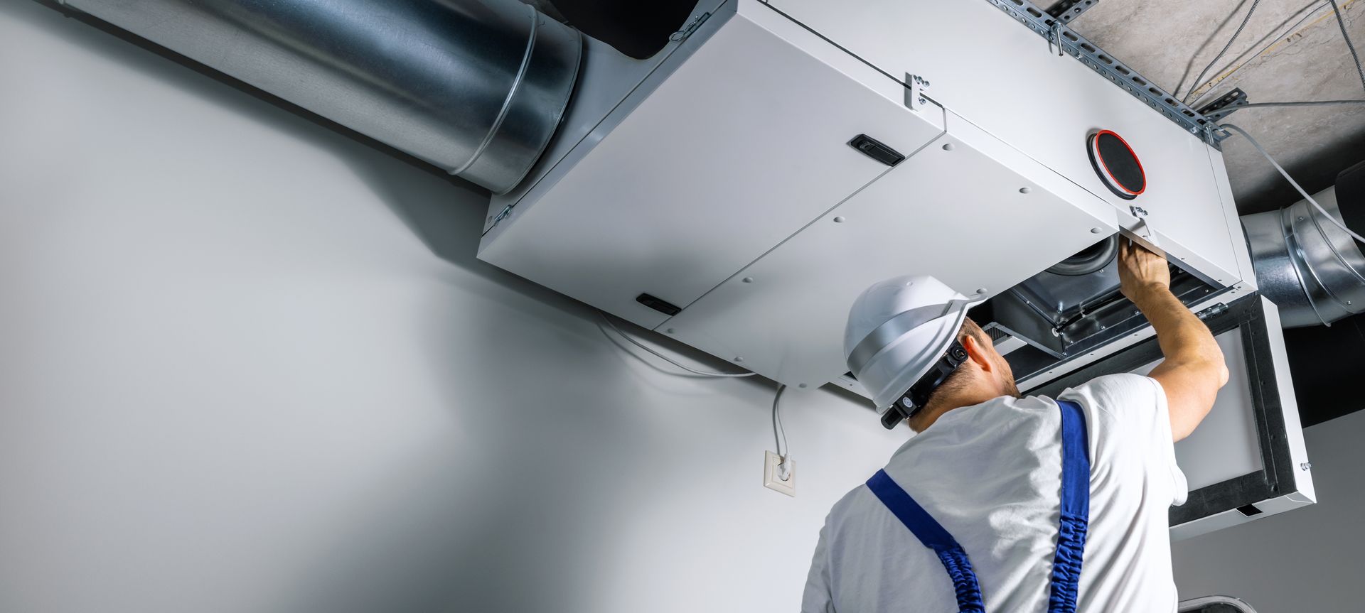 Un technicien portant un casque blanc et une combinaison de travail inspecte une unité de ventilation industrielle CVC fixée au plafond.