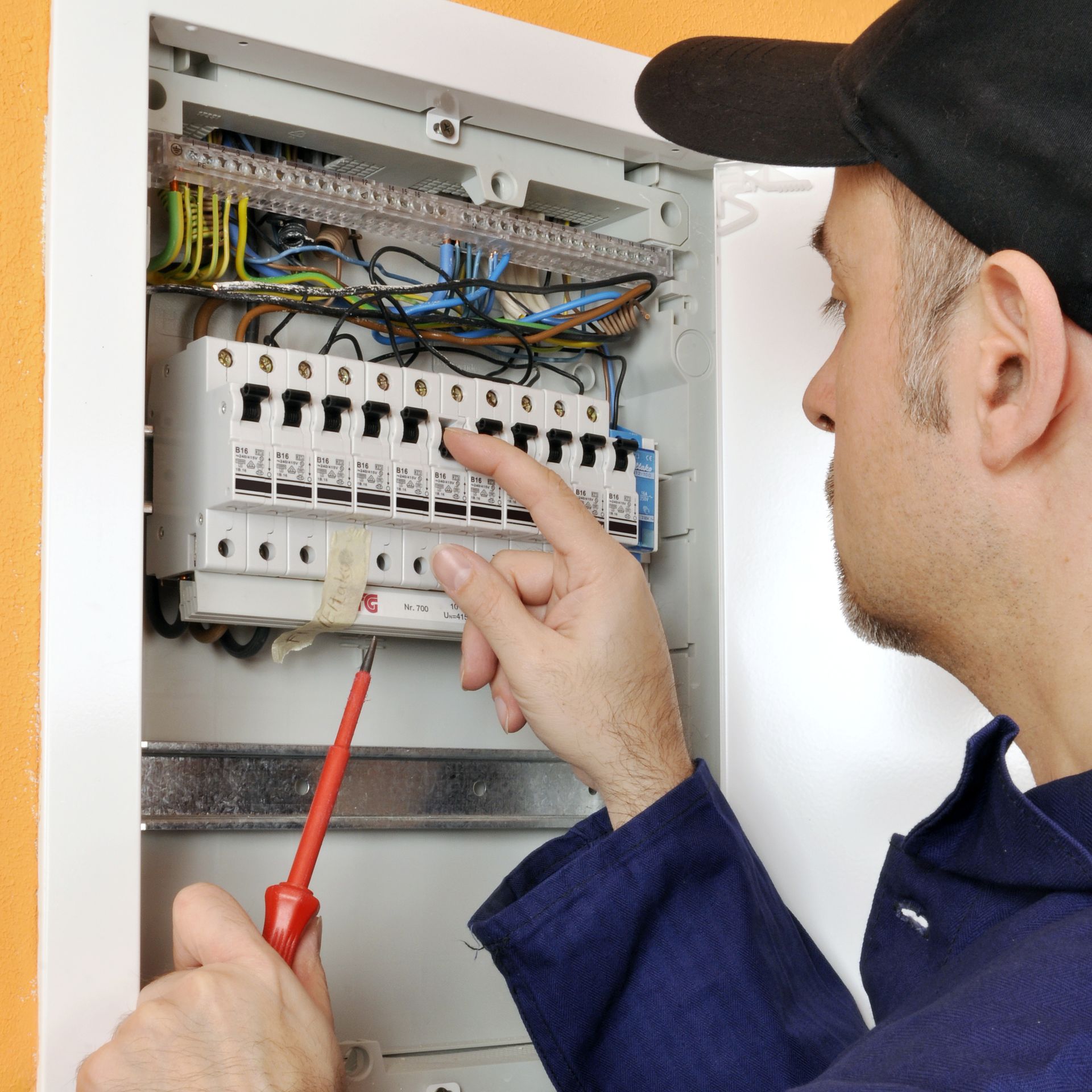 Un électricien en chemise bleue désigne un disjoncteur dans un tableau électrique ouvert tout en tenant un tournevis.