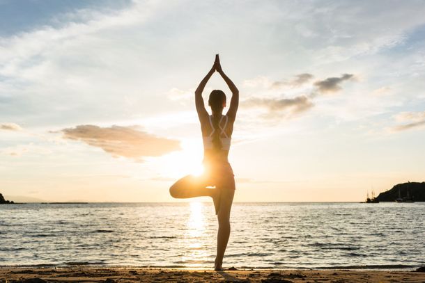 Une personne pratiquant la posture de l'arbre en yoga sur une plage au coucher du soleil, le soleil se reflétant sur l'eau.