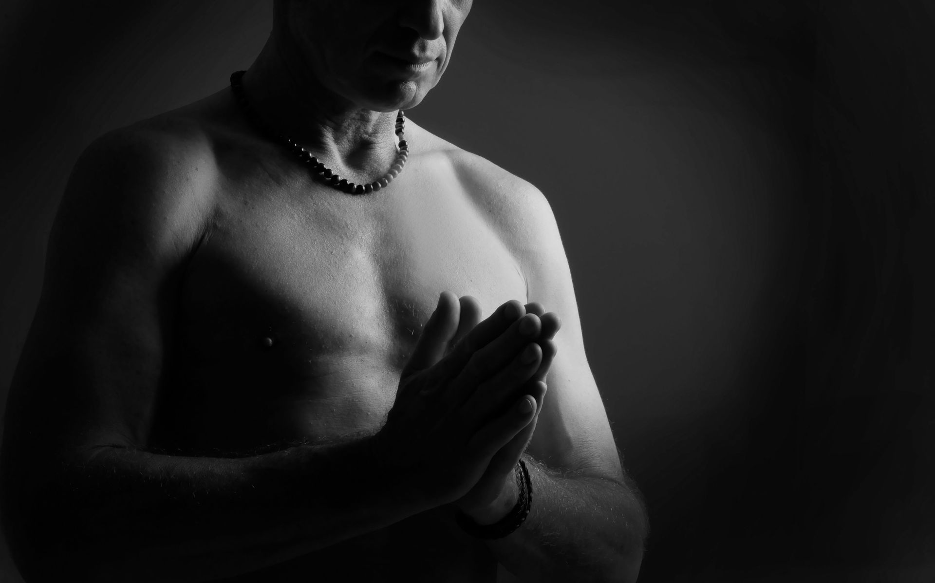 Portrait en noir et blanc d'une personne torse nu, les mains jointes en prière, sur fond sombre.