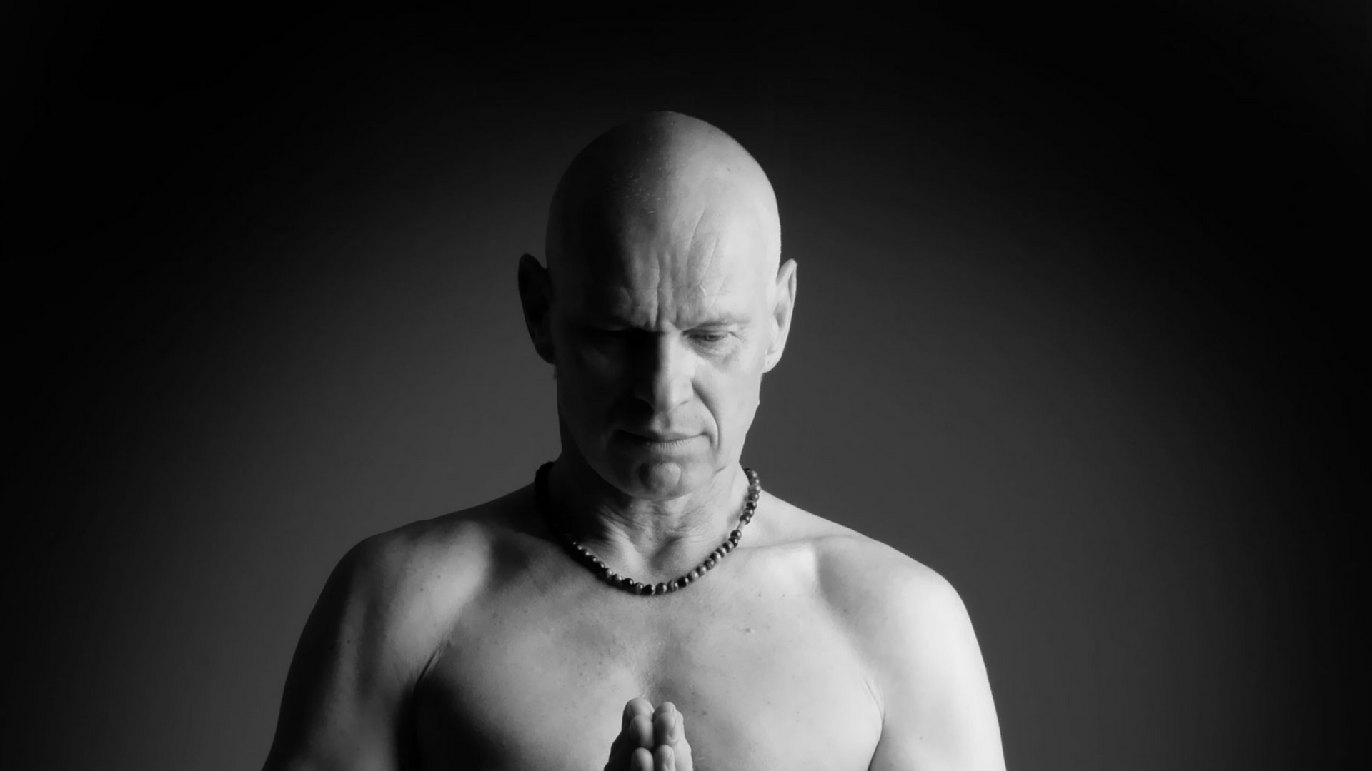 Portrait en noir et blanc d'un homme chauve, les mains jointes en prière, le regard baissé, sur fond sombre.