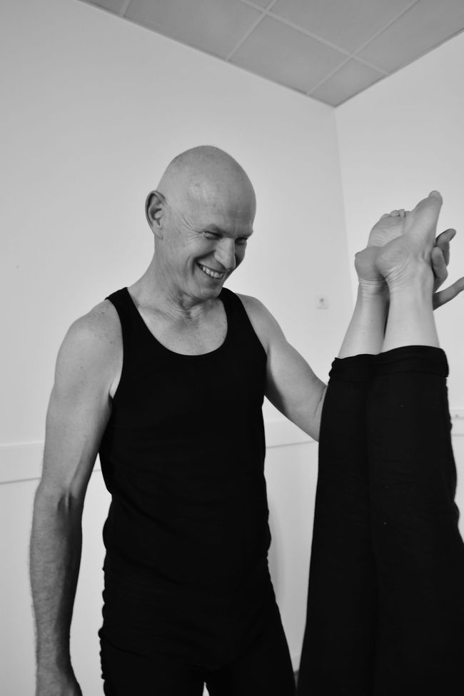 Un homme en débardeur noir aide une personne à faire un étirement vertical des jambes à l'intérieur.