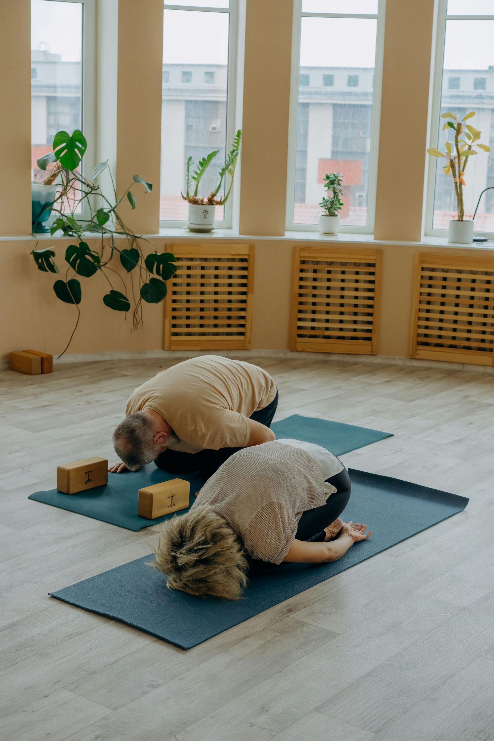 Deux enfants agenouillés sur des tapis de yoga dans un studio lumineux avec des plantes en pot et des appuis de fenêtre en bois.
