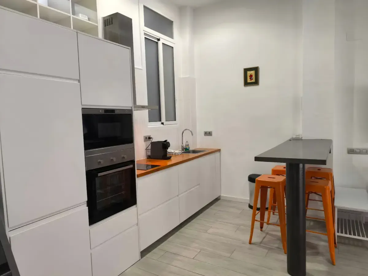 Cocina moderna blanca con horno empotrado, encimera de madera y dos taburetes naranjas junto a una mesa pequeña.
