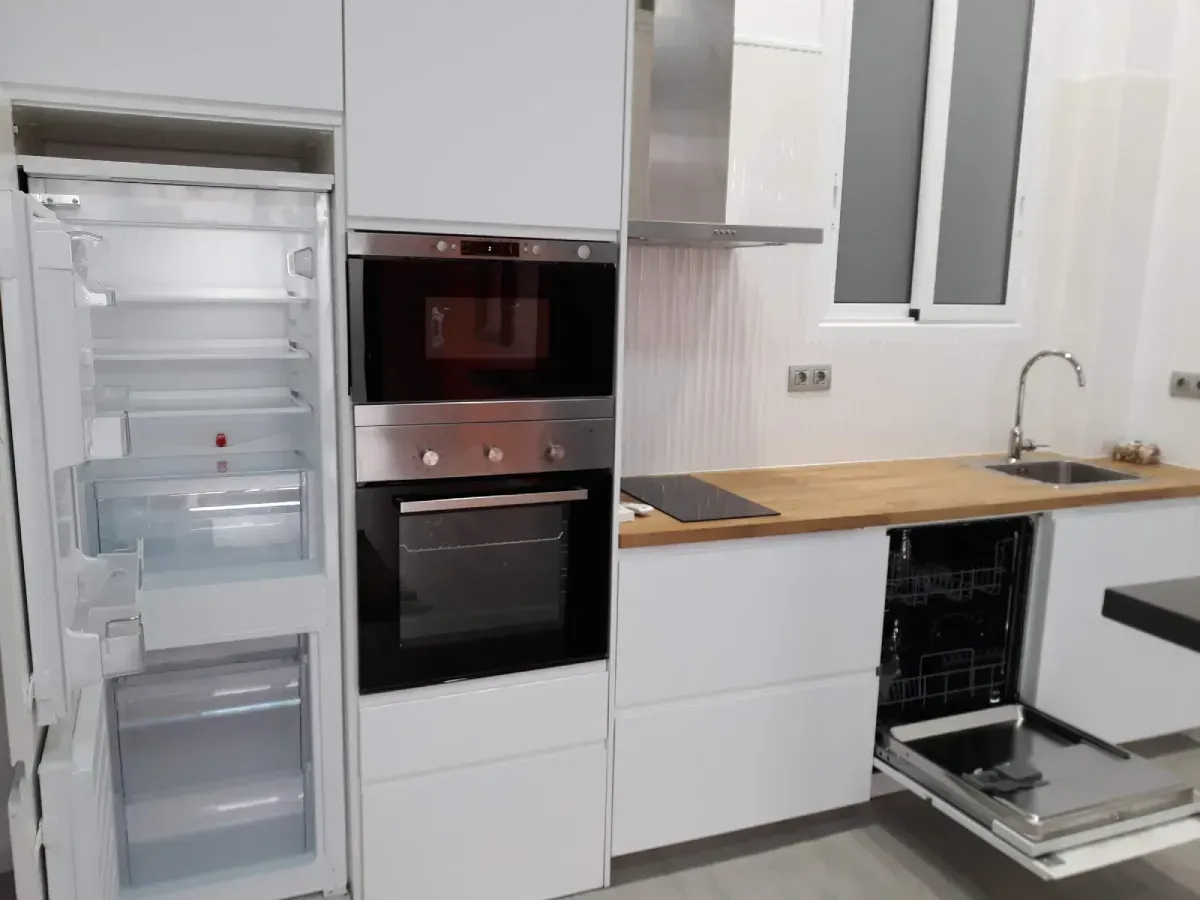 Cocina moderna blanca con horno empotrado, nevera abierta y armarios sin acabado bajo una encimera de madera.
