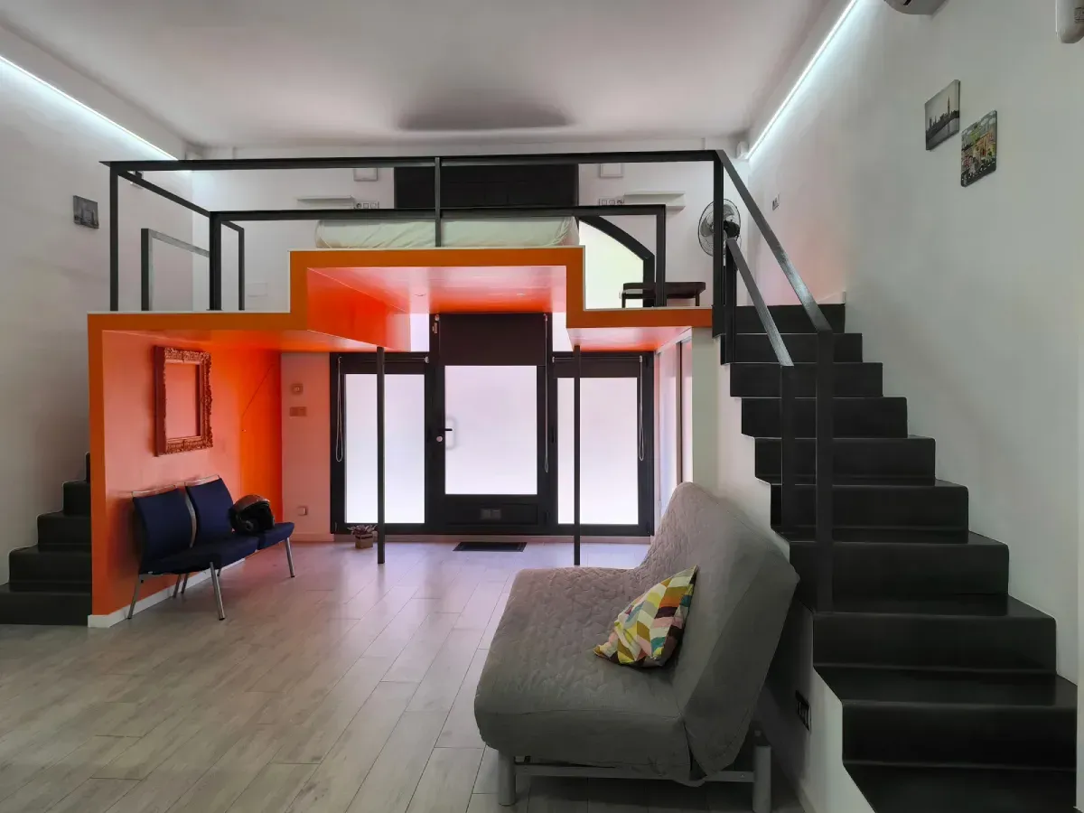 Sala de estar tipo loft moderna con escalera negra, pared decorativa naranja y sofá gris.