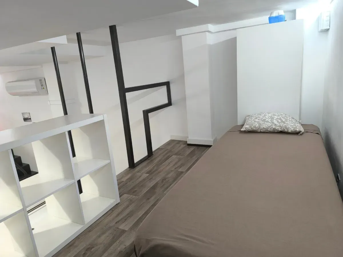 Habitación minimalista con una cama individual, estanterías blancas y suelo de madera gris bajo un techo inclinado.