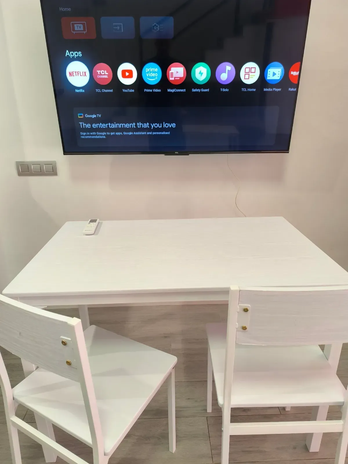 Mesa de comedor blanca con dos sillas en una habitación, frente a un televisor montado en la pared