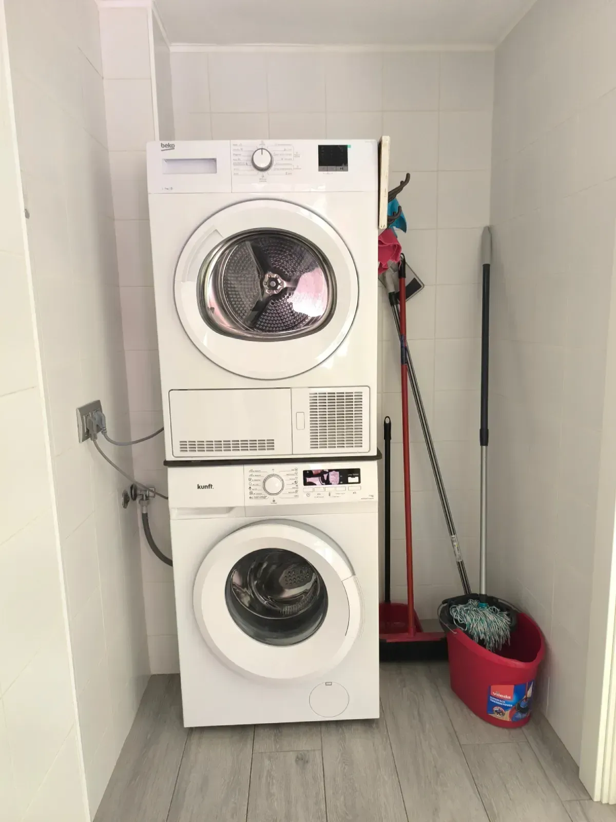 Lavadora y secadora apiladas en un estrecho cuarto de servicio con una fregona y un cubo rojo cerca.