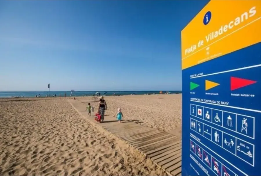 Playa soleada con gente en la arena, cielo azul y un gran cartel azul a la derecha que dice
