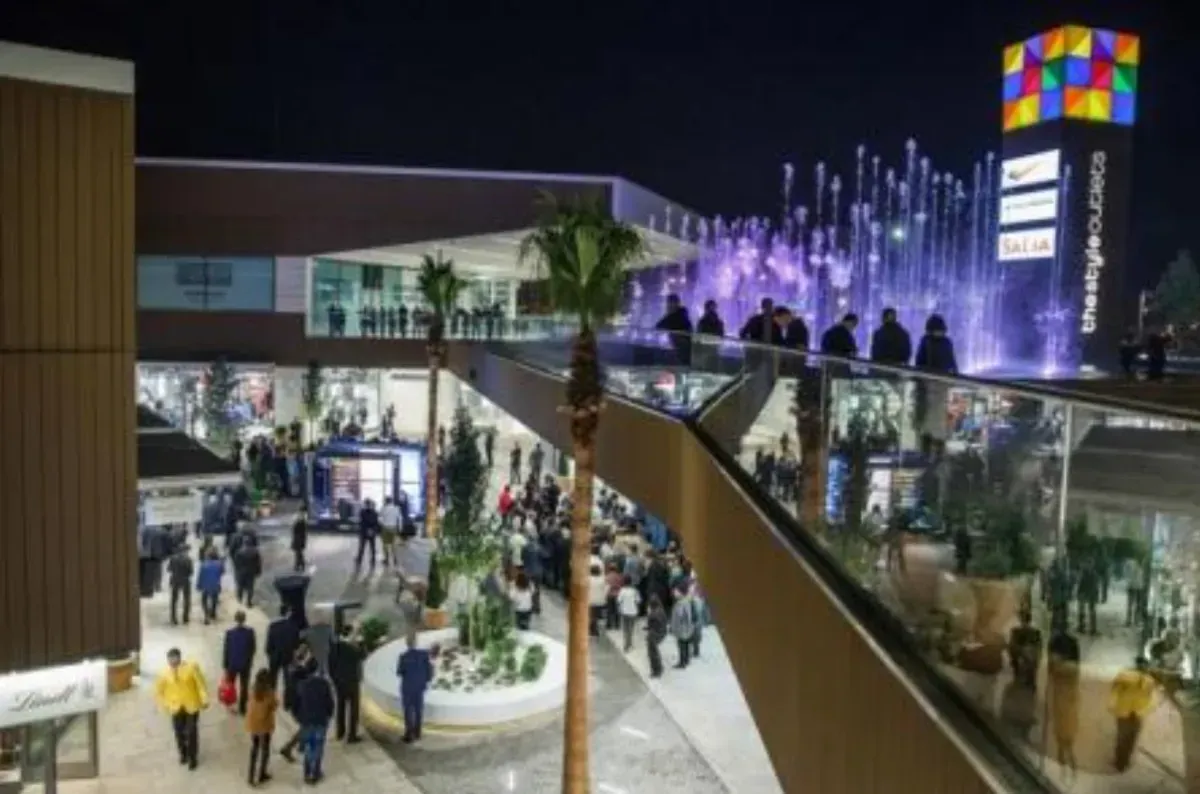 Plaza comercial al aire libre abarrotada de gente por la noche, con luces de fuente moradas y un colorido letrero cúbico.