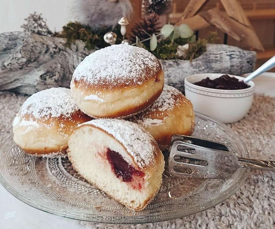 Tisch mit einem Teller Berliner im Vordergrund, einem Schälchen Marmelade daneben und im Hintergrund Weihnachtsdeko bestehend aus einem Wichtel auf einem Deko Ast mit Moos und einem Holz-Tannenbaum