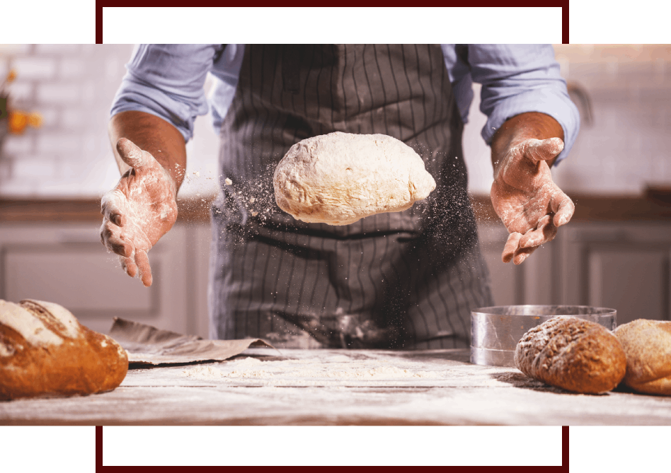 Bäcker an einer Arbeitsplatte, der frisches Brot auf einer Mehlunterlage formt. Rechts und links davon liegen fertige Brote