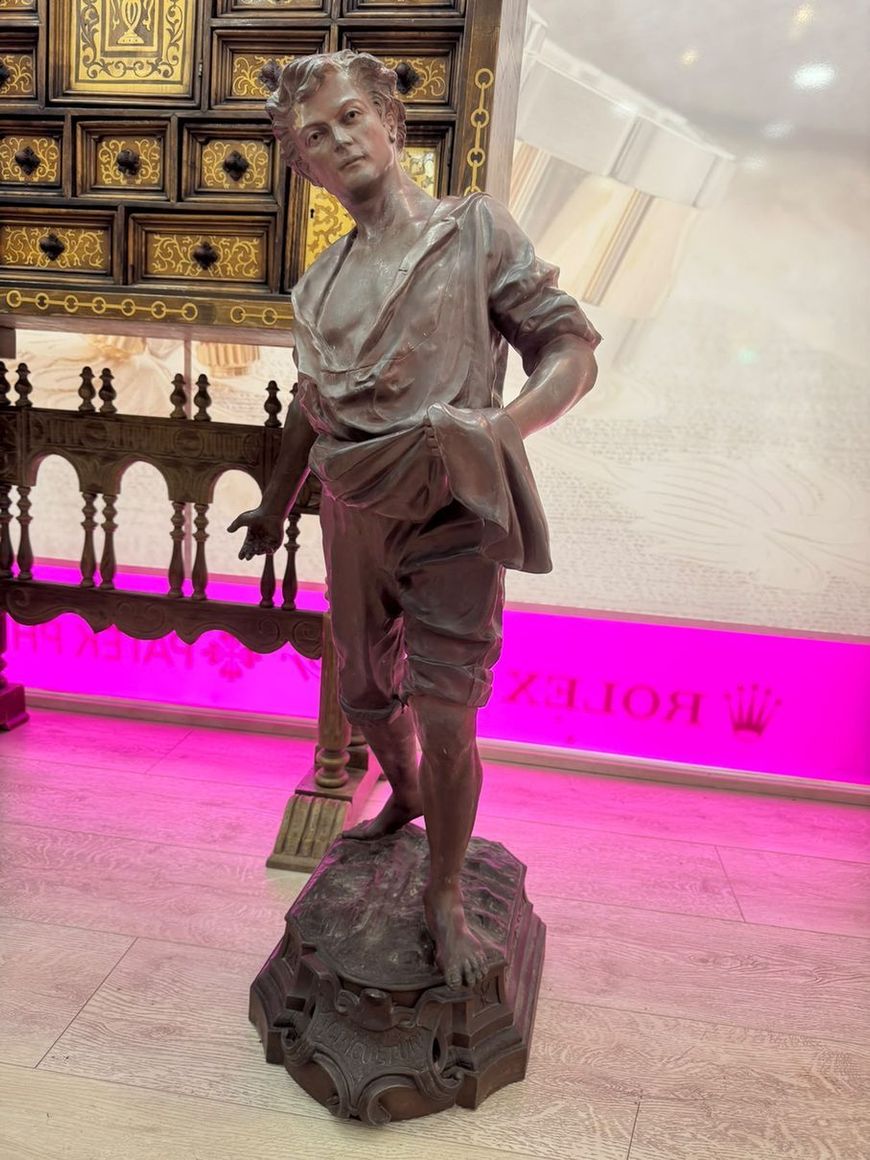Estatua de bronce de un joven músico con un violín, expuesta en interiores sobre un suelo pulido y con un fondo iluminado en rosa.