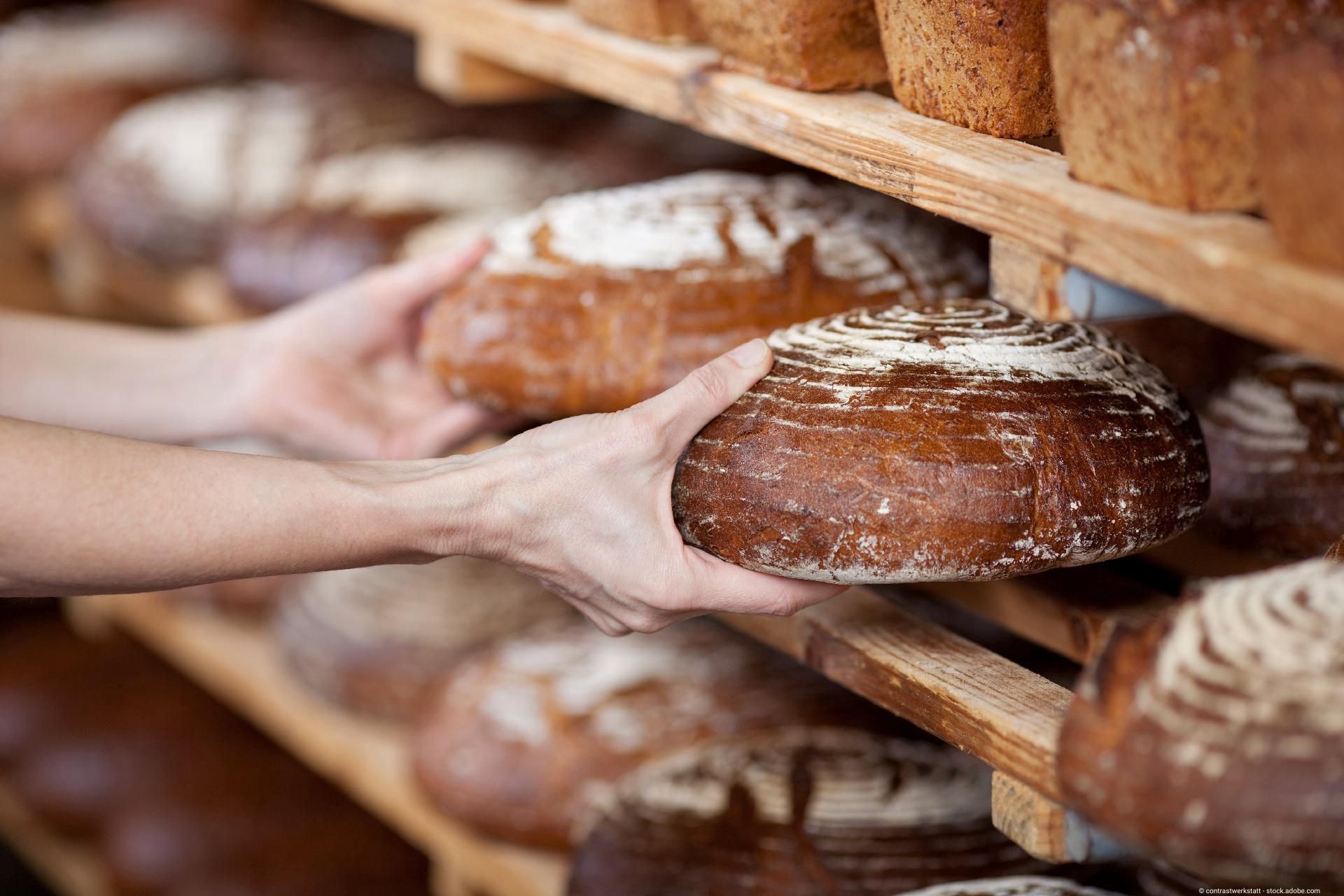 Hände greifen frisch gebackenes Brot aus einem Regal in einer Bäckerei.