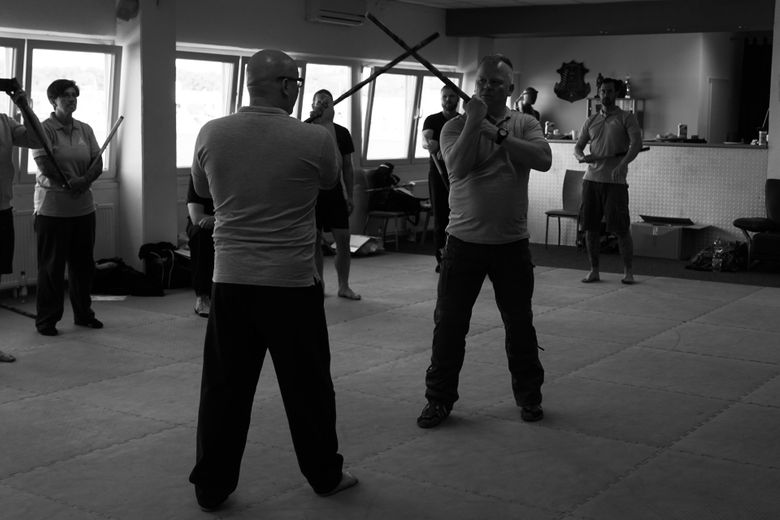 Zwei Kampfsporttrainer demonstrieren den Stockkampf vor einer Gruppe Schüler und Traineranwärter auf der Matte.