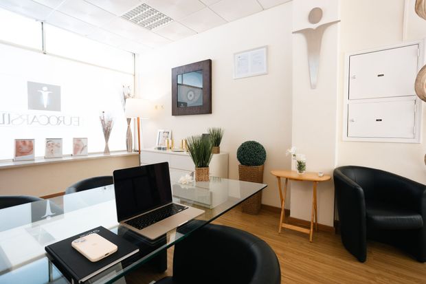 Interior de escritório com mesa de vidro, laptop, cadeiras e vasos de plantas.