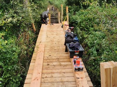 Des ouvriers construisent une passerelle en bois à travers une végétation dense et verte, avec des outils et du bois sur le chemin.